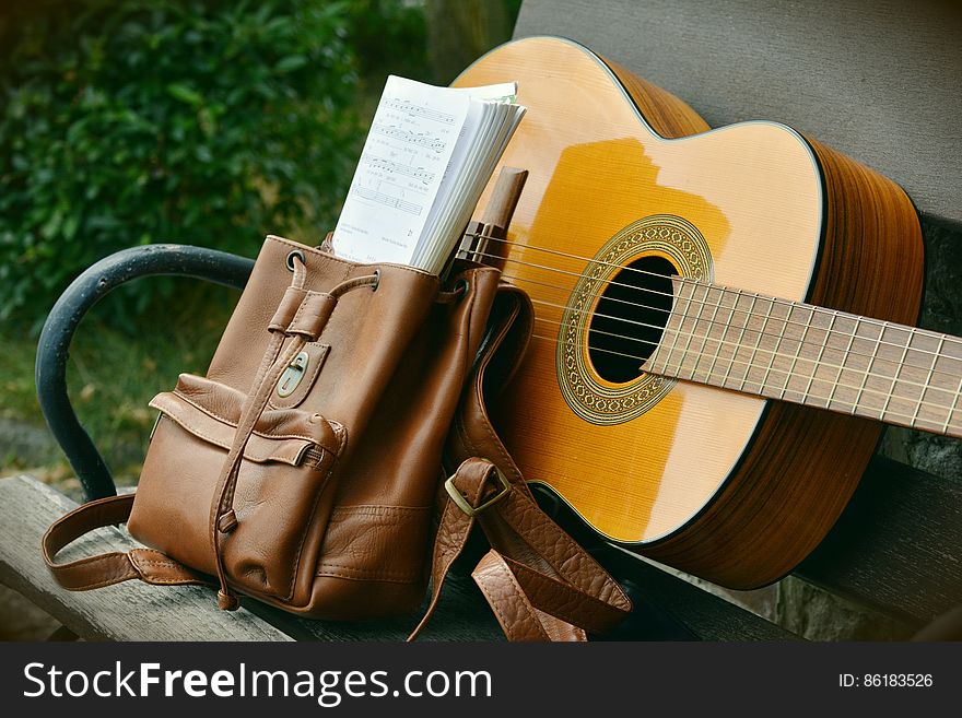 Brown Acoustic Guitar Beside Brown Leather Bucket Backpack on Brown Wooden Bench
