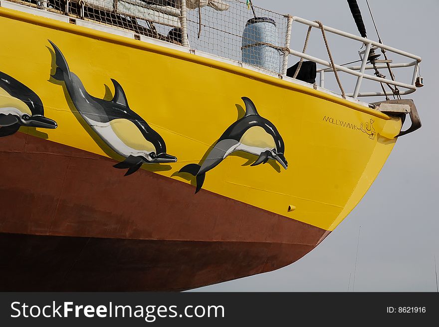 The bow of a sailing boat with three dolphins painted on it in realistic colours. The boat is bright yellow, and her underside is copper coloured. She is ashore, evidently in a boatyard, and we are looking up at the hull. The bow of a sailing boat with three dolphins painted on it in realistic colours. The boat is bright yellow, and her underside is copper coloured. She is ashore, evidently in a boatyard, and we are looking up at the hull.