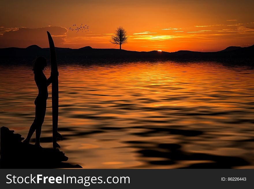 Person Holding a Surfboard Near the Sea on a Setting Sun