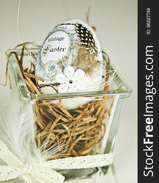 A close up of a traditional Easter egg on straw in a transparent square bowl decorated with white ribbon.