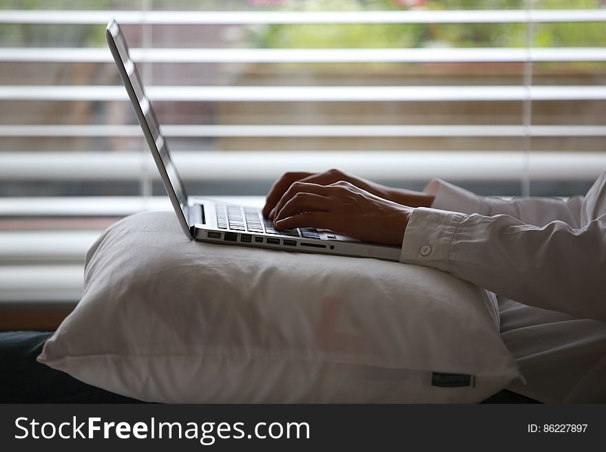 Person Wearing White Dress Shirt Using Silver Laptop on Top of White Throw Pillow