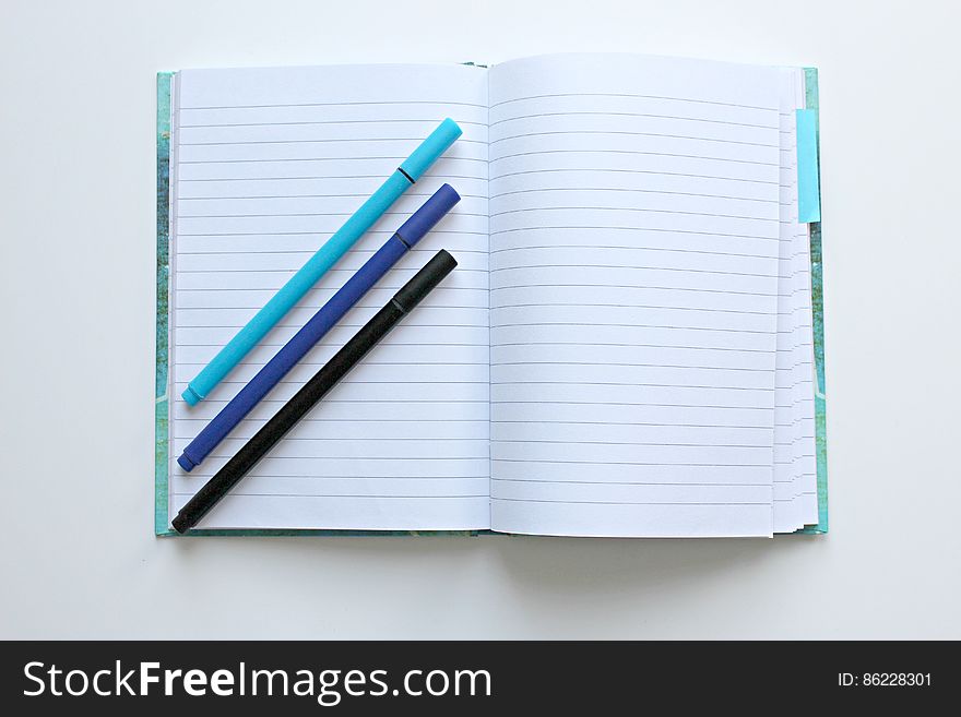 Open lined notebook open with three colored pens lying diagonally across one page, white background. Open lined notebook open with three colored pens lying diagonally across one page, white background.