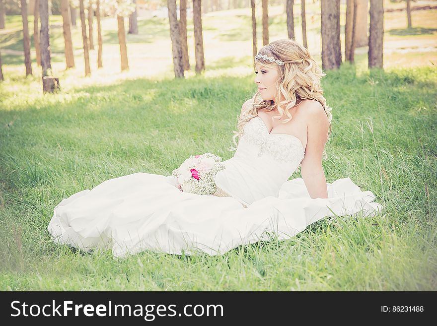 Young blond haired bride sitting in green field with bouquet on sunny day. Young blond haired bride sitting in green field with bouquet on sunny day.