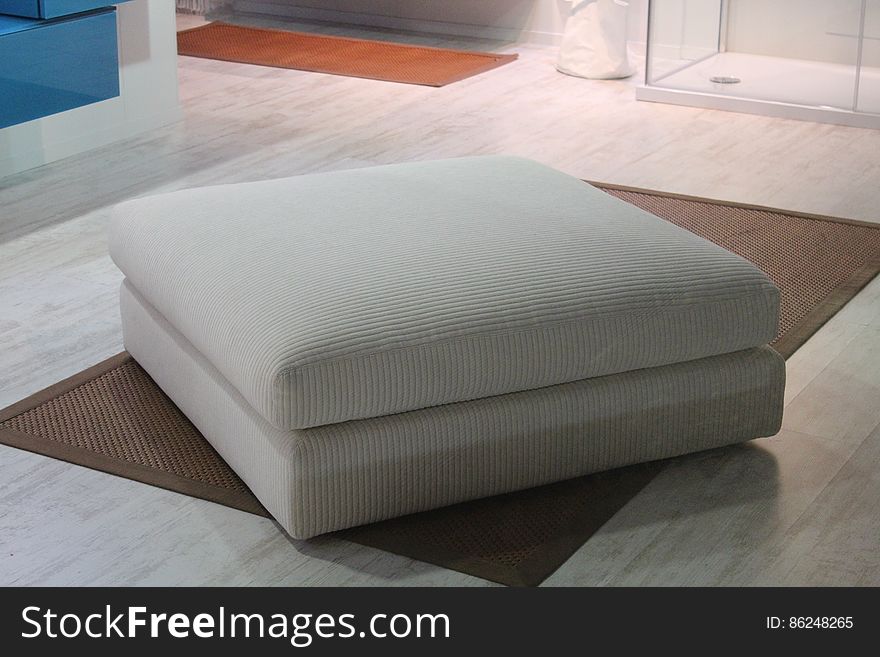 White fabric ottoman on brown area rug inside room of house. White fabric ottoman on brown area rug inside room of house.
