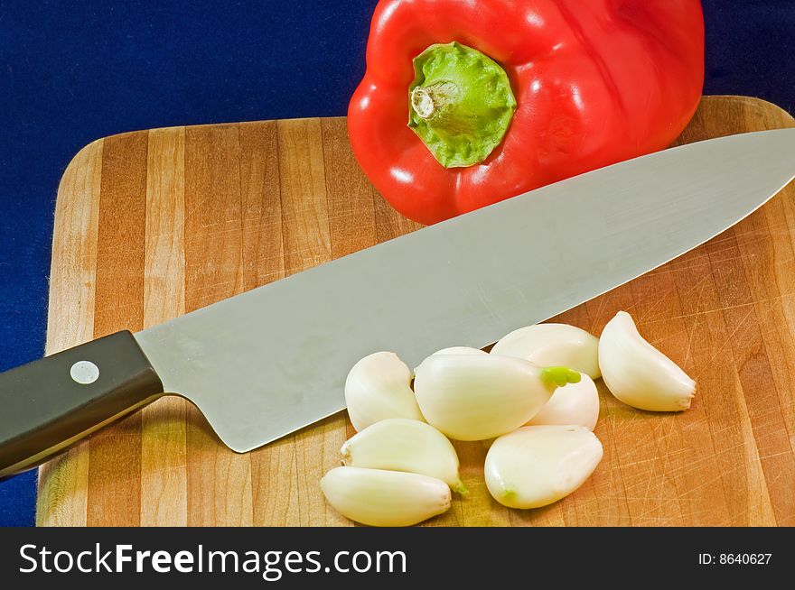 Whole red pepper on a wooden cutting board with garlic cloves and a large kitchen knife. Whole red pepper on a wooden cutting board with garlic cloves and a large kitchen knife.