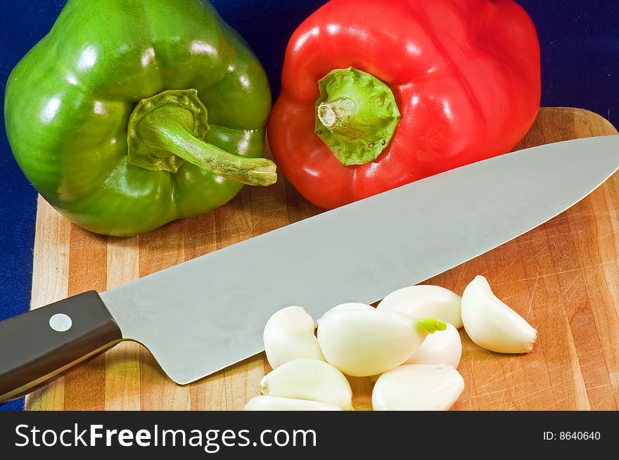 Whole red and green peppers on a wooden cutting board with garlic cloves and a large kitchen knife. Whole red and green peppers on a wooden cutting board with garlic cloves and a large kitchen knife.