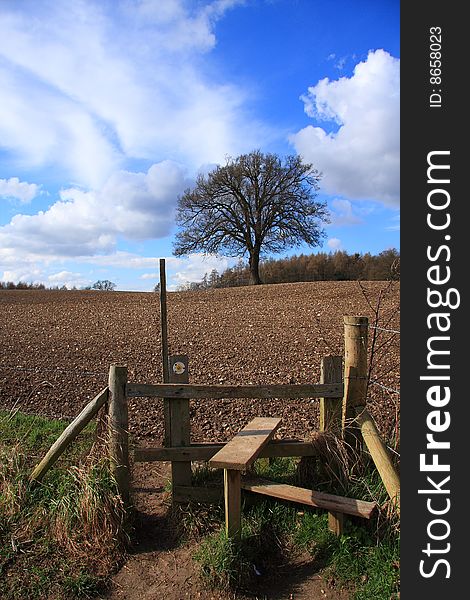 A footpath stile frames the lone tree on the horizon the brown ploughed field contrasts the blue sky. A footpath stile frames the lone tree on the horizon the brown ploughed field contrasts the blue sky