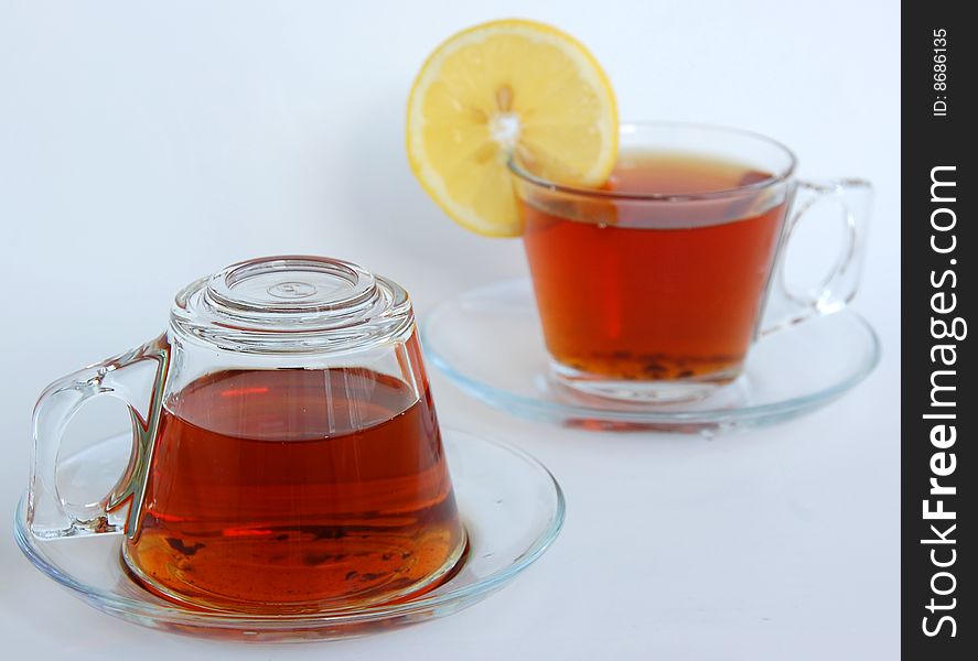 Two full glass teacups of modern design with handle and saucer, one upside-down, the other has a slice of yellow lemon. Two full glass teacups of modern design with handle and saucer, one upside-down, the other has a slice of yellow lemon.