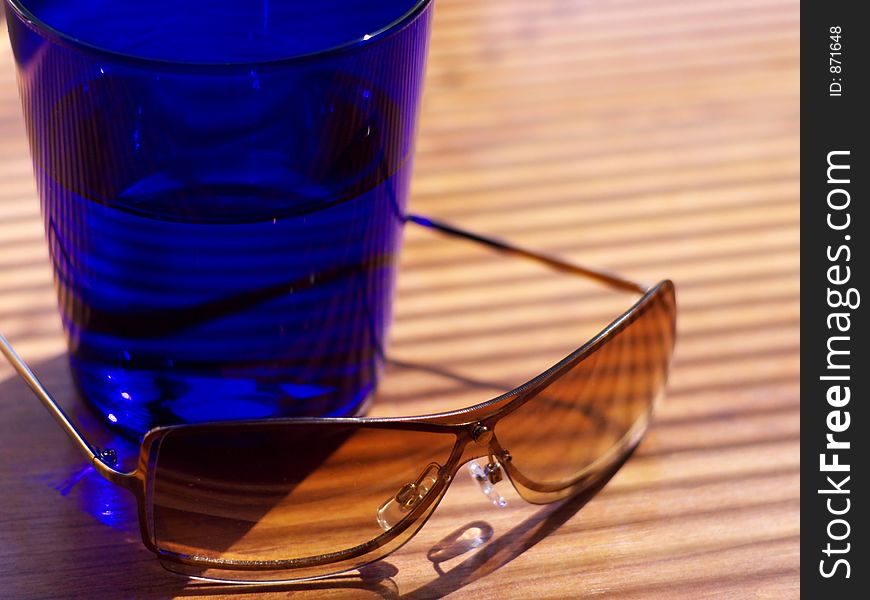 Womens' sunglasses resting next to a dark blue cocktail tumbler in strong light in shallow focus. Womens' sunglasses resting next to a dark blue cocktail tumbler in strong light in shallow focus.