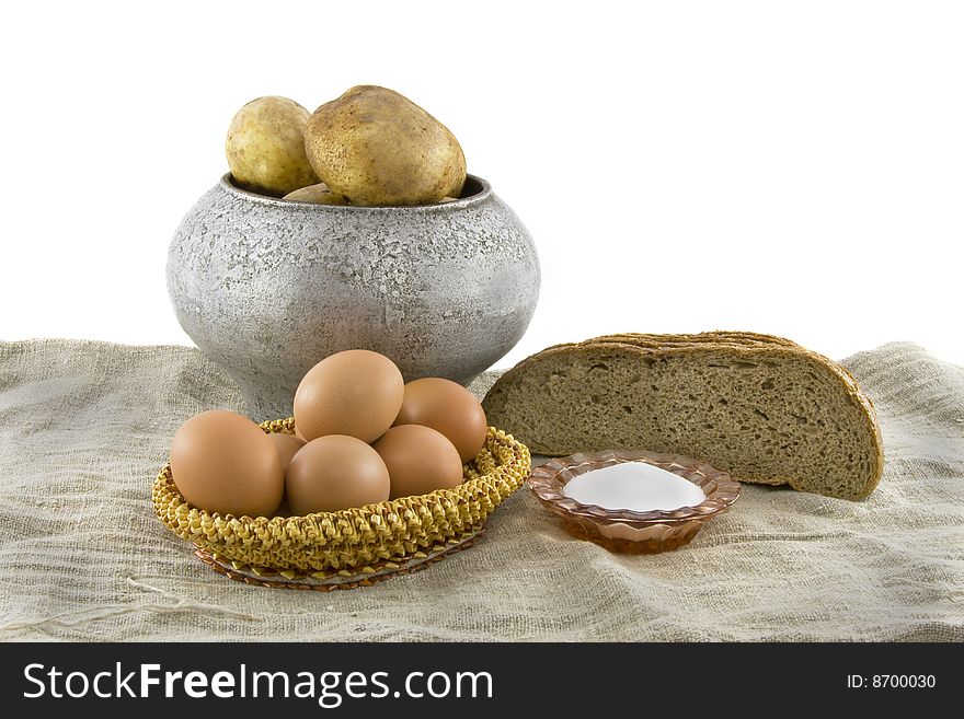 Still-life from vegetarian food. Fresh cucumbers, eggs lying in a wattled small basket, a ripe tomato, fragrant rye bread, a boiled potato in a pig-iron kettle, a saltcellar on a rough fabric