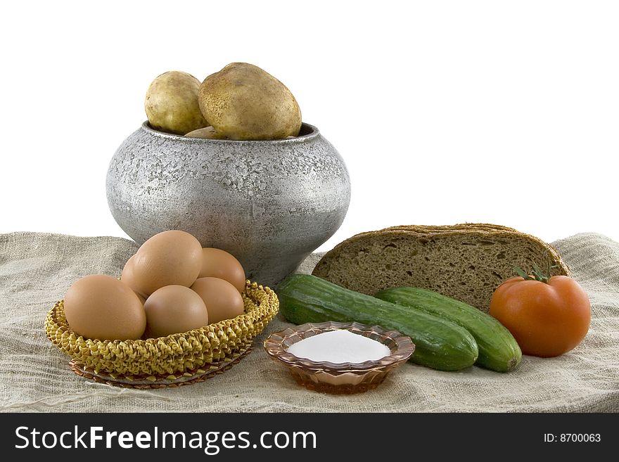 Still-life from vegetarian food. Fresh cucumbers, eggs lying in a wattled small basket, a ripe tomato, fragrant rye bread, a boiled potato in a pig-iron kettle, a saltcellar on a rough fabric