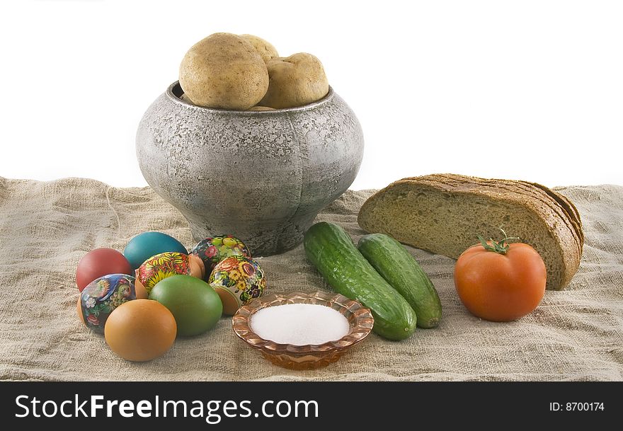 Still-life from vegetarian food. Fresh cucumbers, easter decorated rural eggs, a ripe tomato, fragrant rye bread, a boiled potato in a pig-iron kettle, a saltcellar, lying on sacking rough fabric. Isolated over white background