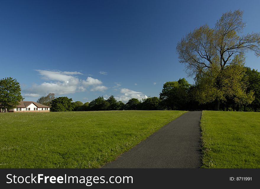 A path stretching away across an empty park with an almost clear blue sky in the background. Space for text in the sky or on the grass.