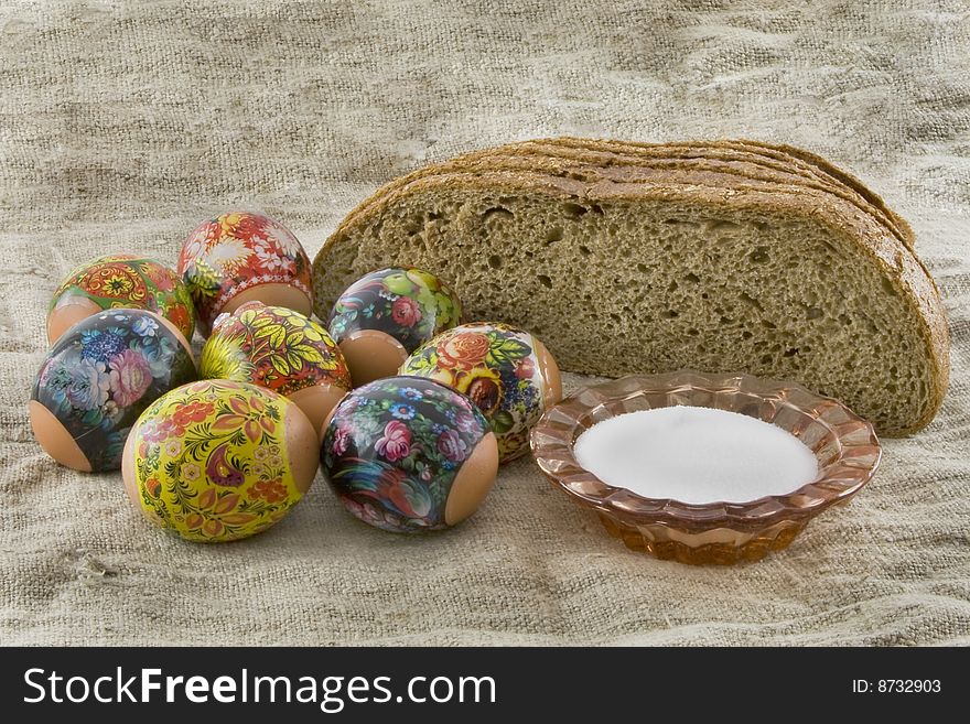 Many fresh easter rural decorated eggs lying in a wattled small basket near to fresh bread and saltcellar on a cloth from a rough fabric. Many fresh easter rural decorated eggs lying in a wattled small basket near to fresh bread and saltcellar on a cloth from a rough fabric