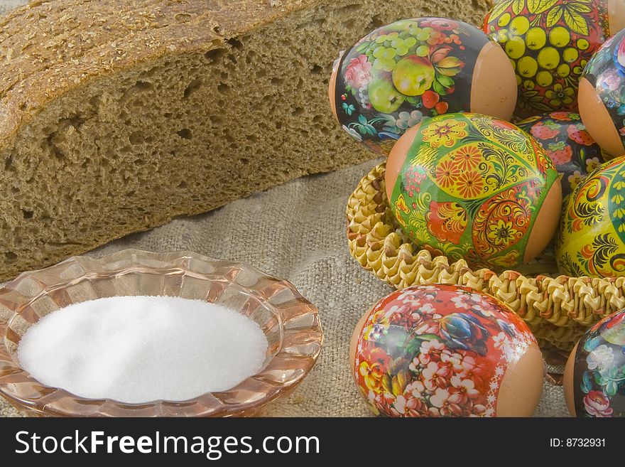 Many fresh easter rural decorated eggs lying in a wattled small basket near to fresh bread and saltcellar on a cloth from a rough fabric