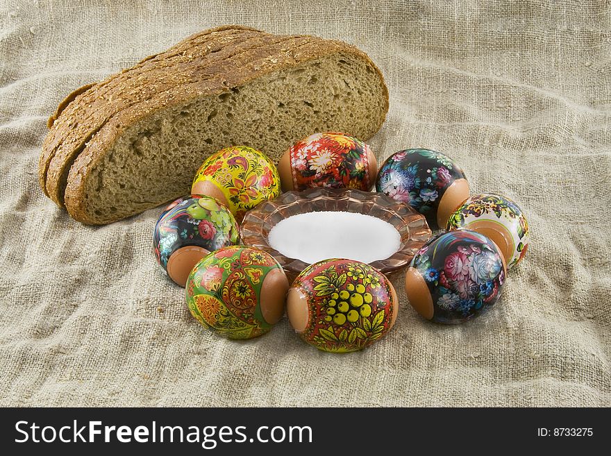 Many fresh easter rural decorated eggs lying in a wattled small basket near to fresh bread and saltcellar on a cloth from a rough fabric