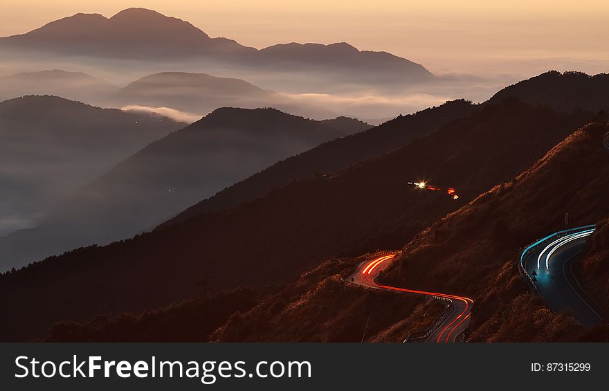 Cars zigzagging along a dark mountain road with headlights and rear lights highlighting the road at dawn producing continuous red and yellow tracks. Cars zigzagging along a dark mountain road with headlights and rear lights highlighting the road at dawn producing continuous red and yellow tracks.