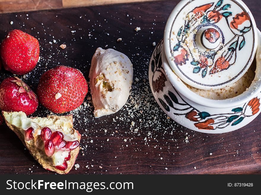 A close up of strawberries, whipped cream, piece of pomegranate and a jar of sugar with crumbs around on a wooden table. A close up of strawberries, whipped cream, piece of pomegranate and a jar of sugar with crumbs around on a wooden table.