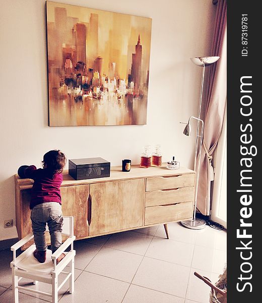Child climbing on chair in living room by cabinet with objects on top.