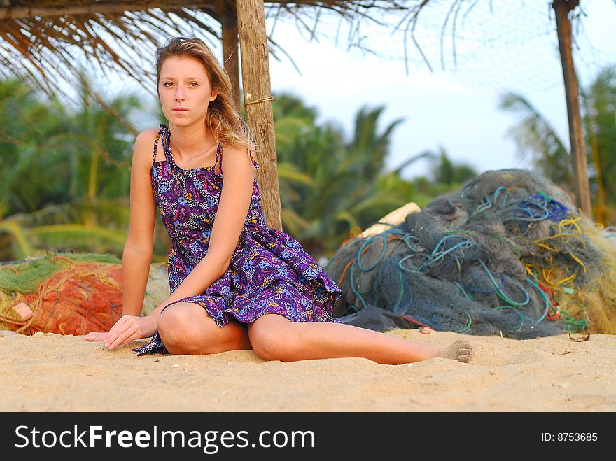 Beautiful young blond girl sitting on tropical beach with fishing nets in background. Beautiful young blond girl sitting on tropical beach with fishing nets in background.