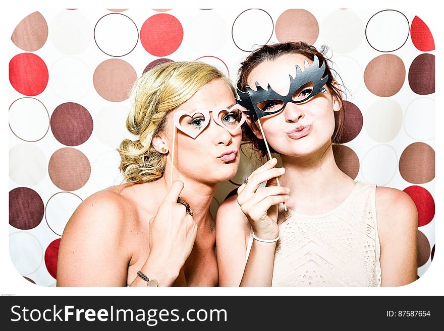 Two Woman Taking Photo in Photobooth Holding Black and Pink Masquerade Mask
