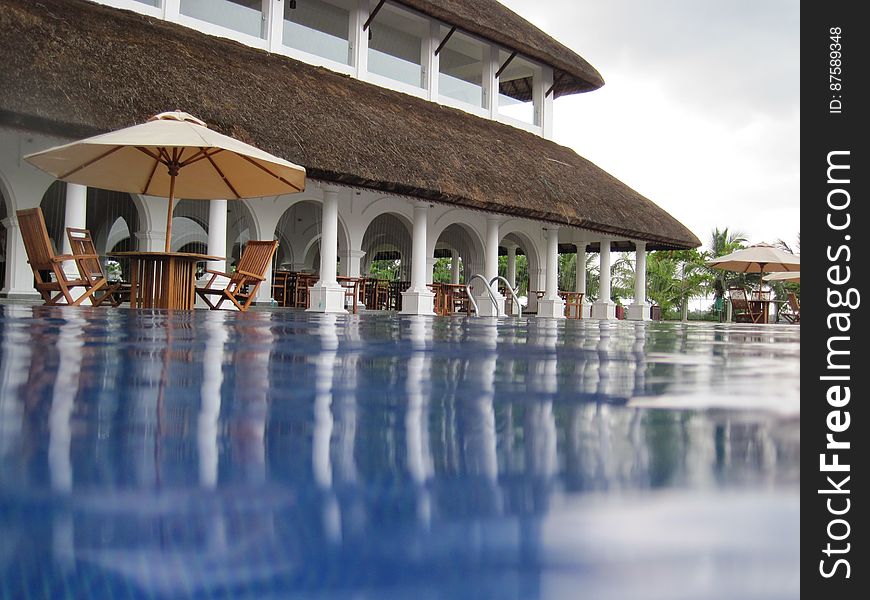 Unusual modern hotel with thatched roof, swimming pool with reflections and covered dining area and next to the pool, table with large umbrella (parasol) and chairs. Unusual modern hotel with thatched roof, swimming pool with reflections and covered dining area and next to the pool, table with large umbrella (parasol) and chairs.