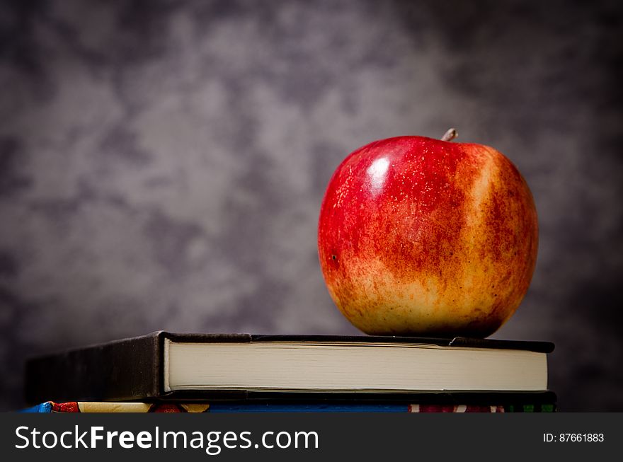 Closeup of rosy red apple placed upon the top of a hardback book, blurred gray background. Closeup of rosy red apple placed upon the top of a hardback book, blurred gray background.