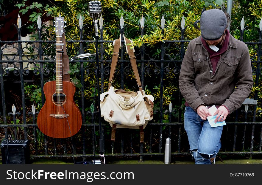 Man Leaning Against Black Steel Fence Beside White And Brown Sling Bag And Brown Acoustic Guitar