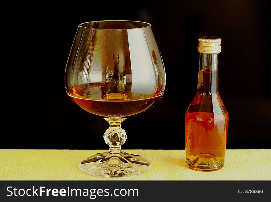 Very small brandy bottle and normal size of glass. Black background. Contrast with light yellow cloth. Very small brandy bottle and normal size of glass. Black background. Contrast with light yellow cloth.