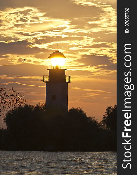 Setting sun seems to lit an old lighthouse while a flock of starlings fly against the orange colored sky. Setting sun seems to lit an old lighthouse while a flock of starlings fly against the orange colored sky.