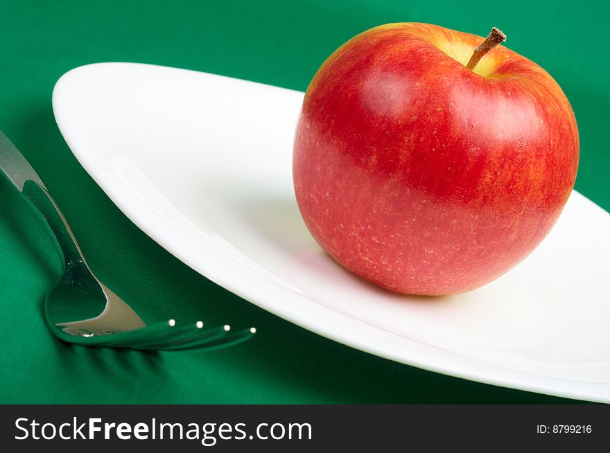 Red apple on white plate and fork on green background. Red apple on white plate and fork on green background