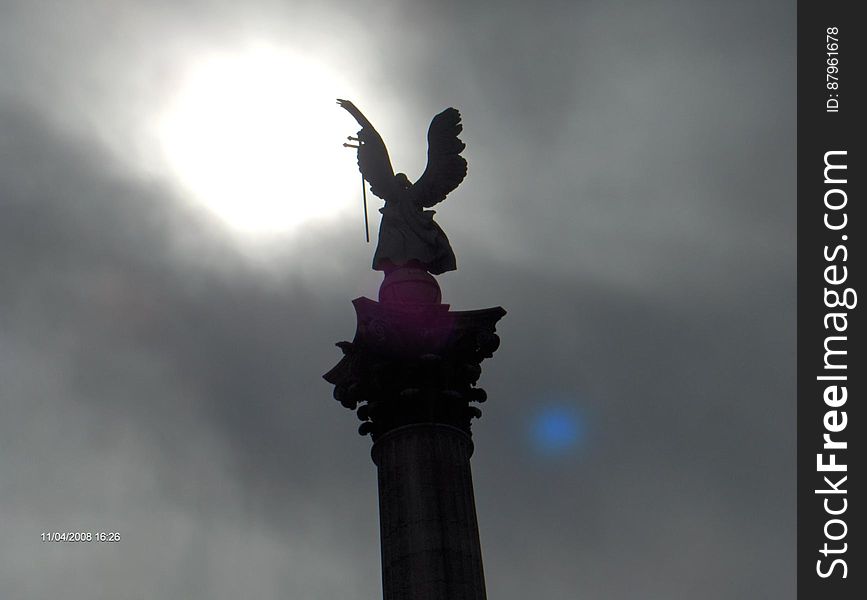 A silhouetted angel statue and the sun breaking through clouds in the background.