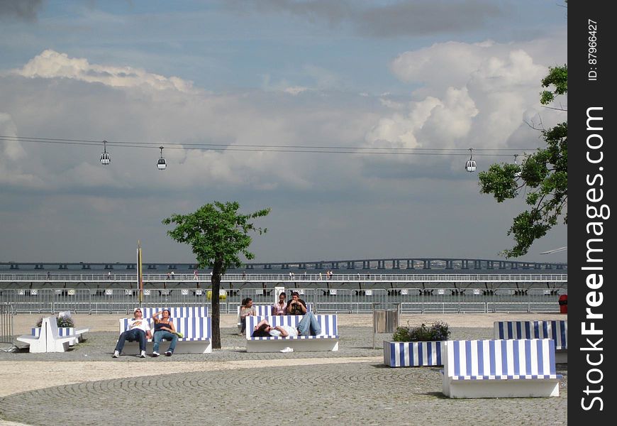 People resting on striped white blue benches in a park and cable car above. People resting on striped white blue benches in a park and cable car above.