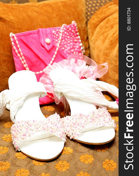 White bridal shoes adorned with pink, white and silver beads with a pink bag and garter in the background lying on brown chair. White bridal shoes adorned with pink, white and silver beads with a pink bag and garter in the background lying on brown chair