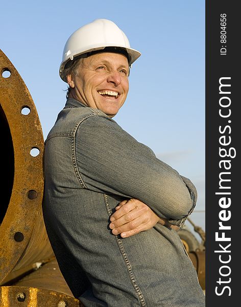 A portrait of a happy smiling construction worker leaning against large metal pipes. A portrait of a happy smiling construction worker leaning against large metal pipes.