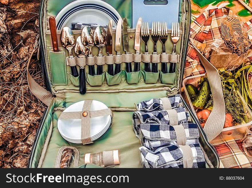 Open picnic bag with cutlery and tableware next to fresh vegetables in countryside.