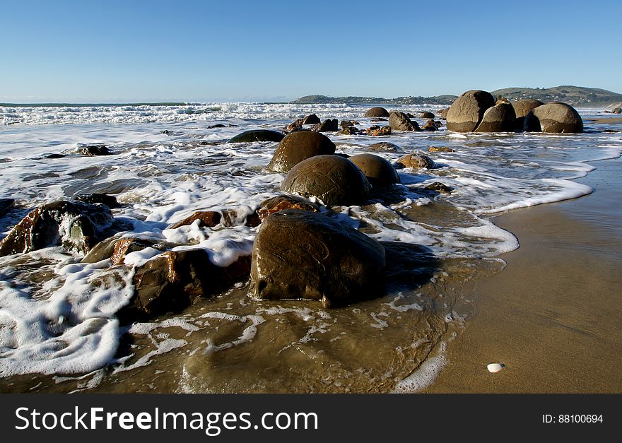 he Moeraki Boulders are unusually large and spherical boulders lying along a stretch of Koekohe Beach on the wave cut Otago coast of New Zealand between Moeraki and Hampden. They occur scattered either as isolated or clusters of boulders within a stretch of beach where they have been protected in a scientific reserve. The erosion by wave action of mudstone, comprising local bedrock and landslides, frequently exposes embedded isolated boulders. These boulders are grey-colored septarian concretions, which have been exhumed from the mudstone enclosing them and concentrated on the beach by coastal erosion. he Moeraki Boulders are unusually large and spherical boulders lying along a stretch of Koekohe Beach on the wave cut Otago coast of New Zealand between Moeraki and Hampden. They occur scattered either as isolated or clusters of boulders within a stretch of beach where they have been protected in a scientific reserve. The erosion by wave action of mudstone, comprising local bedrock and landslides, frequently exposes embedded isolated boulders. These boulders are grey-colored septarian concretions, which have been exhumed from the mudstone enclosing them and concentrated on the beach by coastal erosion
