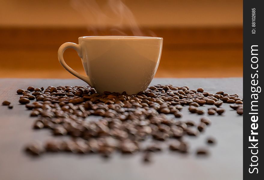 Scattered coffee beans on table with steaming hot cup in background. Scattered coffee beans on table with steaming hot cup in background.