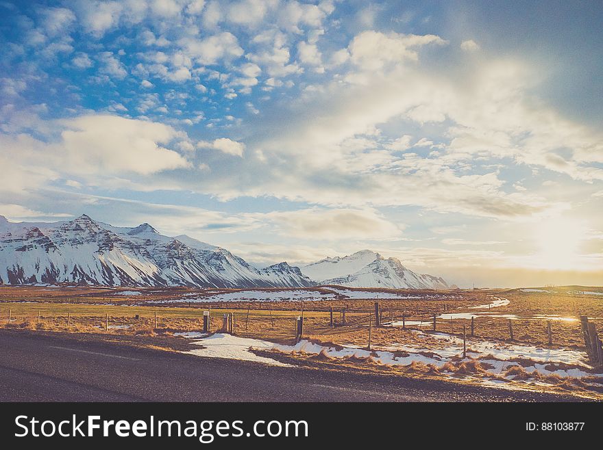 Snow covered mountain range next to country field with empty road on sunny day. Snow covered mountain range next to country field with empty road on sunny day.