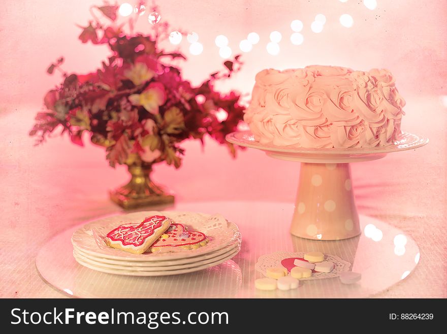 Cake on serving dish with plate of heart shaped cookies next to floral arrangement with pink background. Cake on serving dish with plate of heart shaped cookies next to floral arrangement with pink background.