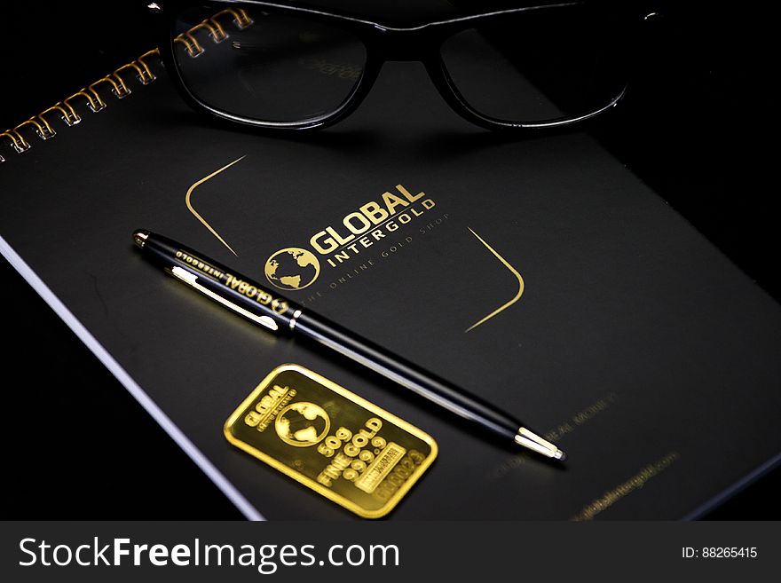 Still life with notebook stamped with Global Intergold logo and text with pen, eyeglasses and gold ingot. Still life with notebook stamped with Global Intergold logo and text with pen, eyeglasses and gold ingot.