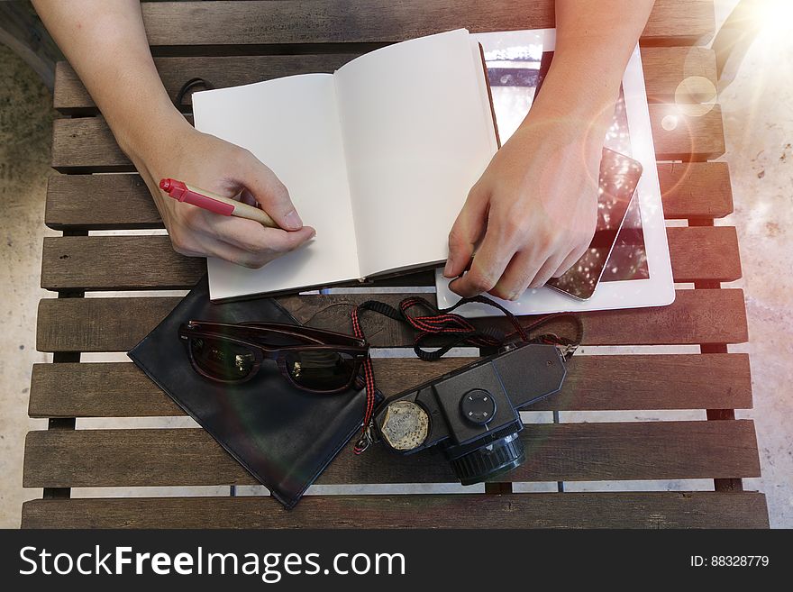 Two disembodied hands, one holding pen writing in a notebook placed on a slatted chair or table surrounded by camera, sun glasses, mobile phone and black case. Two disembodied hands, one holding pen writing in a notebook placed on a slatted chair or table surrounded by camera, sun glasses, mobile phone and black case.