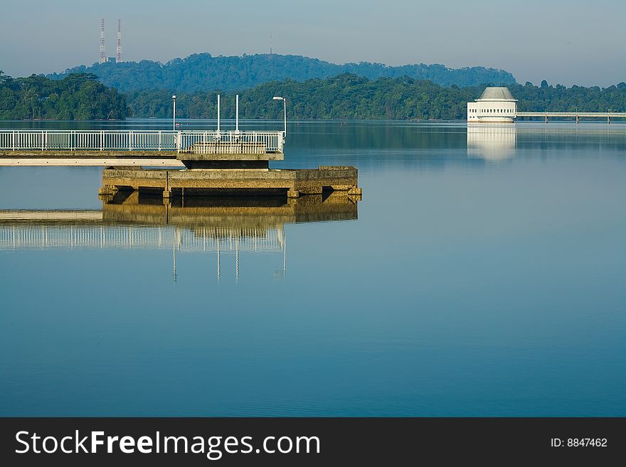 Relflected on the calm water are landmarks of the Upper Peirce Reservoir of Singapore. Iron towers on the Bukit Timah hill, the white water monitoring building, and a jetty are iconic objects of the erservoir.