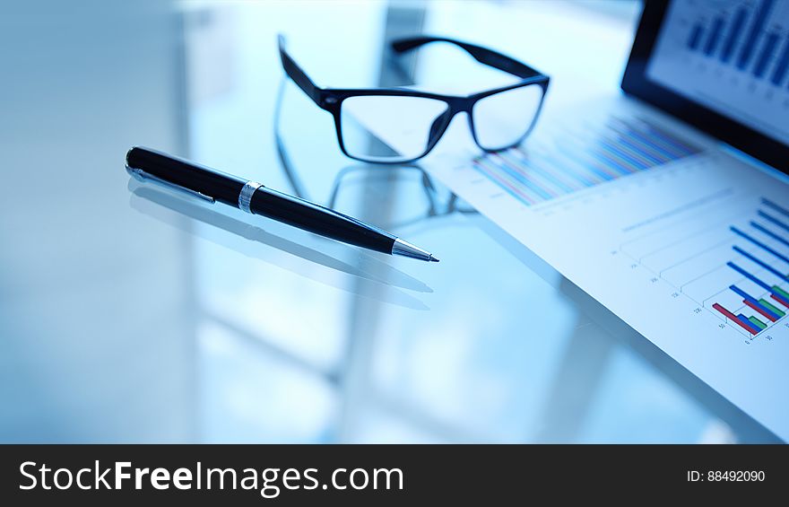 Glasses and pen on blue toned table next to data on business documents and computer screen. Glasses and pen on blue toned table next to data on business documents and computer screen.