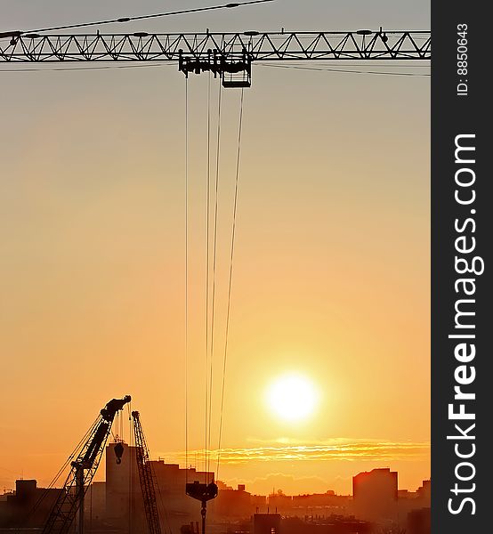 Crane silhouette in upper part of frame with cords hanging down from it and town buildings in the horizone, orange sun in the middle. Crane silhouette in upper part of frame with cords hanging down from it and town buildings in the horizone, orange sun in the middle