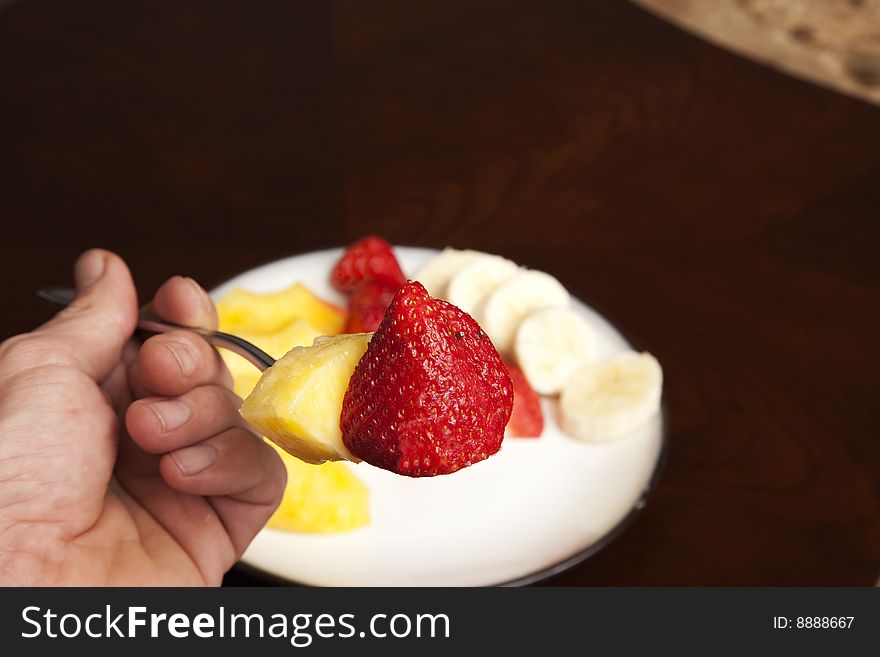 A strawberry and pineapple slice on a fork with a plate of fruit in the background. A strawberry and pineapple slice on a fork with a plate of fruit in the background.