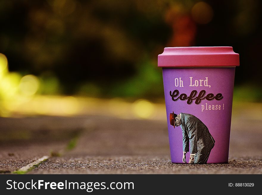 Bed of coffee beans with selective focus on a purple colored mug of carry out coffee inscribed with text "Oh Lord, Coffee please!" blurred background.