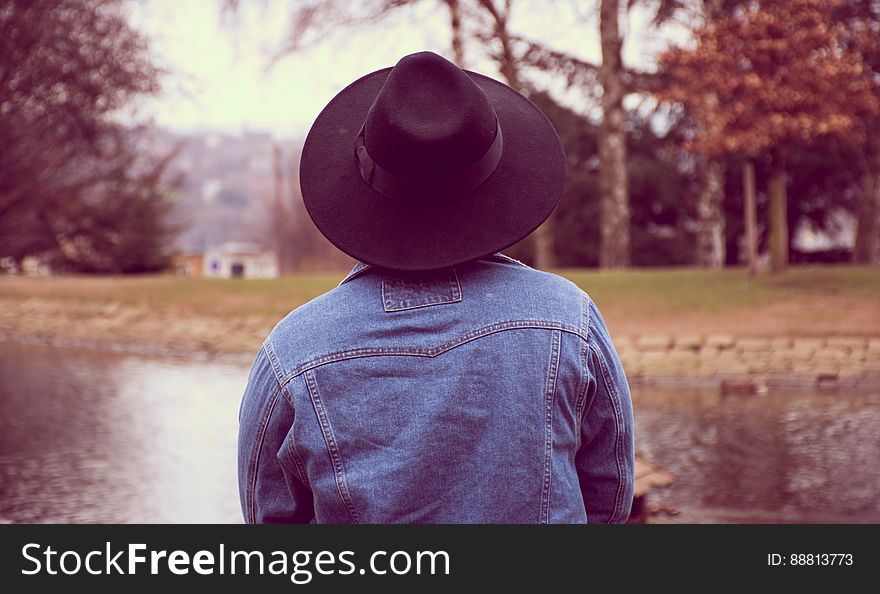 A person wearing a denim jacket and a fedora by a pond in a park. A person wearing a denim jacket and a fedora by a pond in a park.