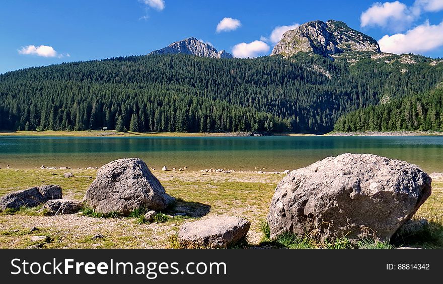 Large rocks on the shore of a lake on the far side of which is a dense fir forest, mountain peaks, blue sky and cloud background. Large rocks on the shore of a lake on the far side of which is a dense fir forest, mountain peaks, blue sky and cloud background.