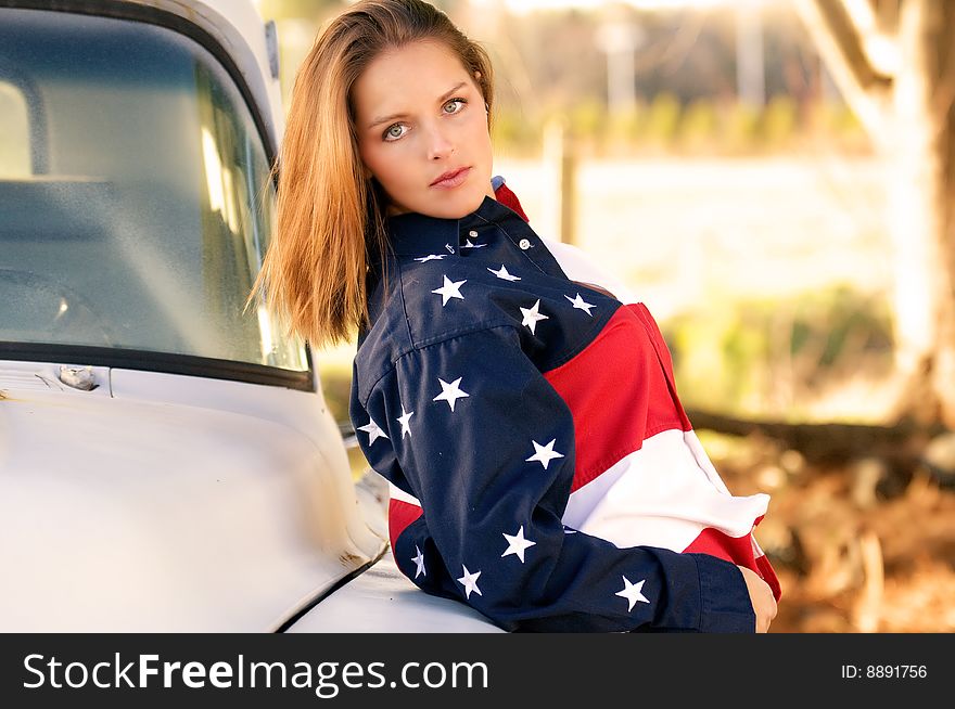 Attractive country girl in American flag shirt leans back against an old truck in rural america. Attractive country girl in American flag shirt leans back against an old truck in rural america